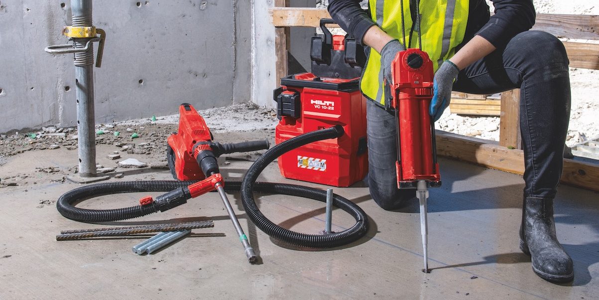 Construction worker using Hilti SafeSet installation method with HDE 500-22 cordless adhesive dispenser and red HIT-CR 500 cartridge holder for HIT-HY 200 adhesives. VC 140-2-22 "vac-pack" vacuum, TE 60-22 cordless rotary hammer